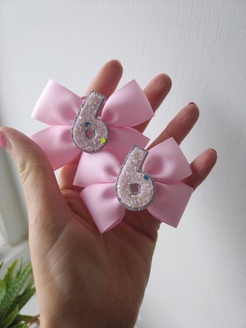 May include: Two pink hair bows with silver glitter number sixes in the centre. The bows are made of satin ribbon and have a sparkly finish.