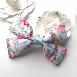 Op de afbeelding: Een witte en blauwe strik met een Peter Rabbit-print. De strik heeft een lichtblauw midden en is versierd met roze bloemen en groene bladeren. De tekst "Peter Rabbit" wordt herhaald in de hele print.