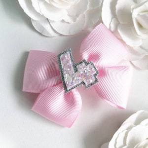 Könnte beinhalten: Eine rosa Schleife mit einer silbernen Glitzerzahl vier in der Mitte. Die Schleife ist aus Satinband gefertigt und hat eine glänzende Oberfläche.