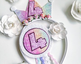 May include: A white headband with a rainbow glitter crown and a circle with the number "4" in pink glitter. The headband is decorated with white flowers and colourful ribbons.