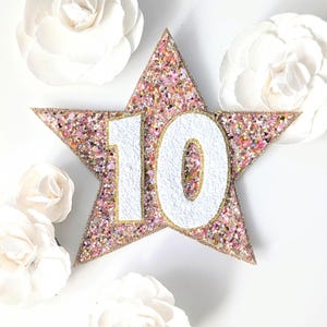 Puede incluir: Una decoración en forma de estrella rosa brillante con el número 10 en blanco. La estrella tiene un borde dorado y está cubierta de purpurina rosa, dorada e iridiscente. Flores de papel blancas en el fondo.