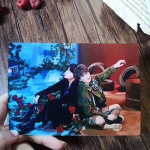 May include: A painting of two people sitting on a floor with a blue and red background. The person on the left is wearing a black outfit and the person on the right is wearing a green jacket and jeans. There are red roses in the foreground.