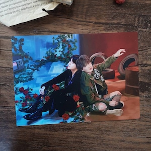 May include: Two men are sitting together in a room with a blue and red background. One man is wearing a black suit and the other is wearing a green jacket. There are red roses in the foreground.