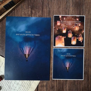 Puede incluir: Dos tarjetas imprimibles con un fondo de cielo nocturno azul y un globo aerostático. El texto en las tarjetas dice "00:00 y vas a ser feliz".