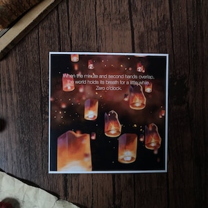 May include: A print with a quote about time and a dark background with many orange and yellow lanterns floating in the air. The quote reads: "When the minute and second hands overlap, the world holds its breath for a little while. Zero o'clock."
