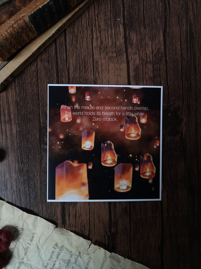 May include: A print with a quote about time and a dark background with many orange and yellow lanterns floating in the air. The quote reads: "When the minute and second hands overlap, the world holds its breath for a little while. Zero o'clock."