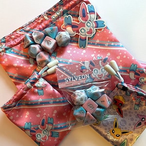 Può includere: Due sacchetti a cordoncino con motivo rosa e blu con un adesivo Sylveon e un sacchetto di dadi blu e rosa. Il sacchetto dei dadi riporta la scritta "Sylveon Dice Set".