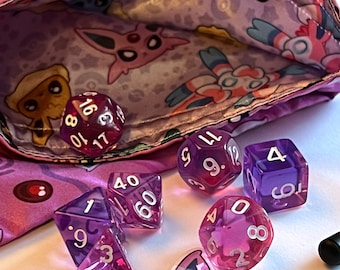 Op de afbeelding: Een paarse stoffen tas met een Pokemon-printontwerp, open om een ​​set van zes paarse en heldere dobbelstenen met witte nummers te onthullen. De dobbelstenen zijn een d4, d6, d8, d10, d12 en d20.