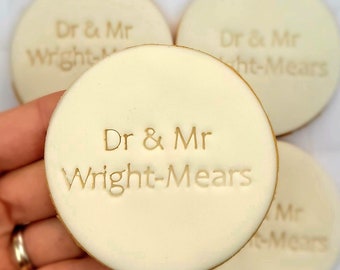 Op de afbeelding: Vier ronde, witte koekjes met gouden letters. De koekjes zijn versierd met de tekst "Dr & Mr Wright-Mears".