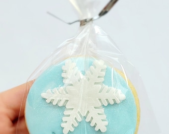 Op de afbeelding: Een lichtblauwe suikerkoek versierd met een witte sneeuwvlok en de naam "Marley" in blauwe glazuur. De koek is verpakt in een doorzichtige plastic zak.