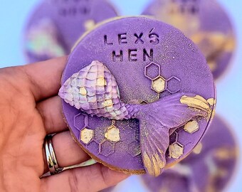 Op de afbeelding: Paarse en gouden gedecoreerde koekjes met een zeemeerminstaartontwerp en de tekst "Lex's Hen" erop.