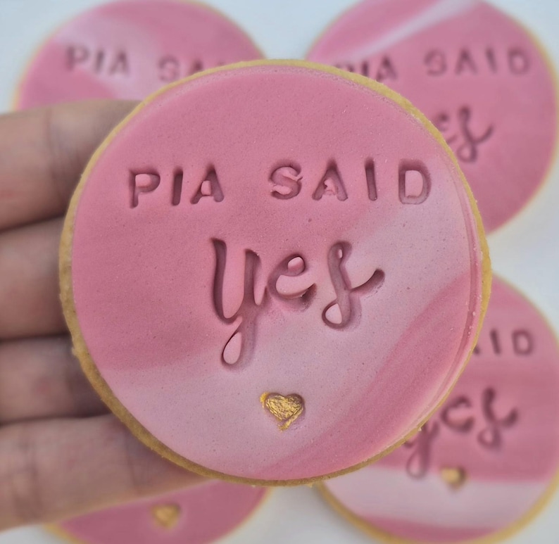 Puede incluir: Una galleta redonda con una cubierta de fondant rosa marmolado que presenta el texto en relieve PIA SAID yes y un peque&ntilde;o coraz&oacute;n dorado. La galleta tiene un borde marr&oacute;n claro y es uno de varios dulces circulares similares con el dise&ntilde;o.