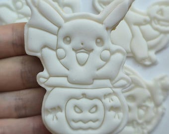 Op de afbeelding: Een wit koekje in de vorm van Pikachu die uit een pompoenemmer komt. Het personage heeft puntige oren, ronde wangen en een glimlachende uitdrukking. De pompoen heeft een uitgesneden lantaarngezicht met kleine vleermuizen aan de zijkanten.