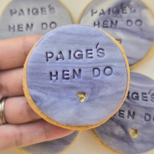 Puede incluir: Una galleta redonda cubierta con glaseado liso de color morado claro jaspeado. El texto "PAIGE'S HEN DO" est&aacute; grabado en el centro, con una peque&ntilde;a forma de coraz&oacute;n rellena de oro situada justo debajo de las letras en el glaseado.