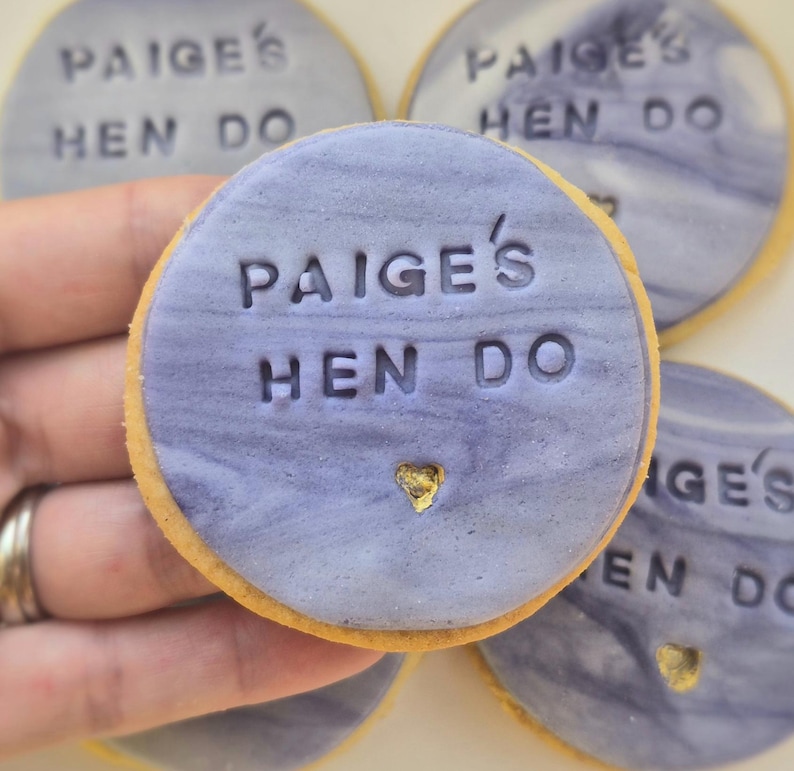 Puede incluir: Una galleta redonda cubierta con glaseado liso de color morado claro jaspeado. El texto "PAIGE'S HEN DO" est&aacute; grabado en el centro, con una peque&ntilde;a forma de coraz&oacute;n rellena de oro situada justo debajo de las letras en el glaseado.
