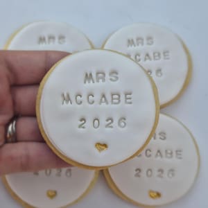 Puede incluir: Cuatro galletas de az&uacute;car redondas con glaseado blanco, cada una grabada con el texto MRS MCCABE 2026 y un peque&ntilde;o coraz&oacute;n dorado. Las galletas tienen un borde dorado claro y est&aacute;n dispuestas en grupo, con una sostenida en primer plano.