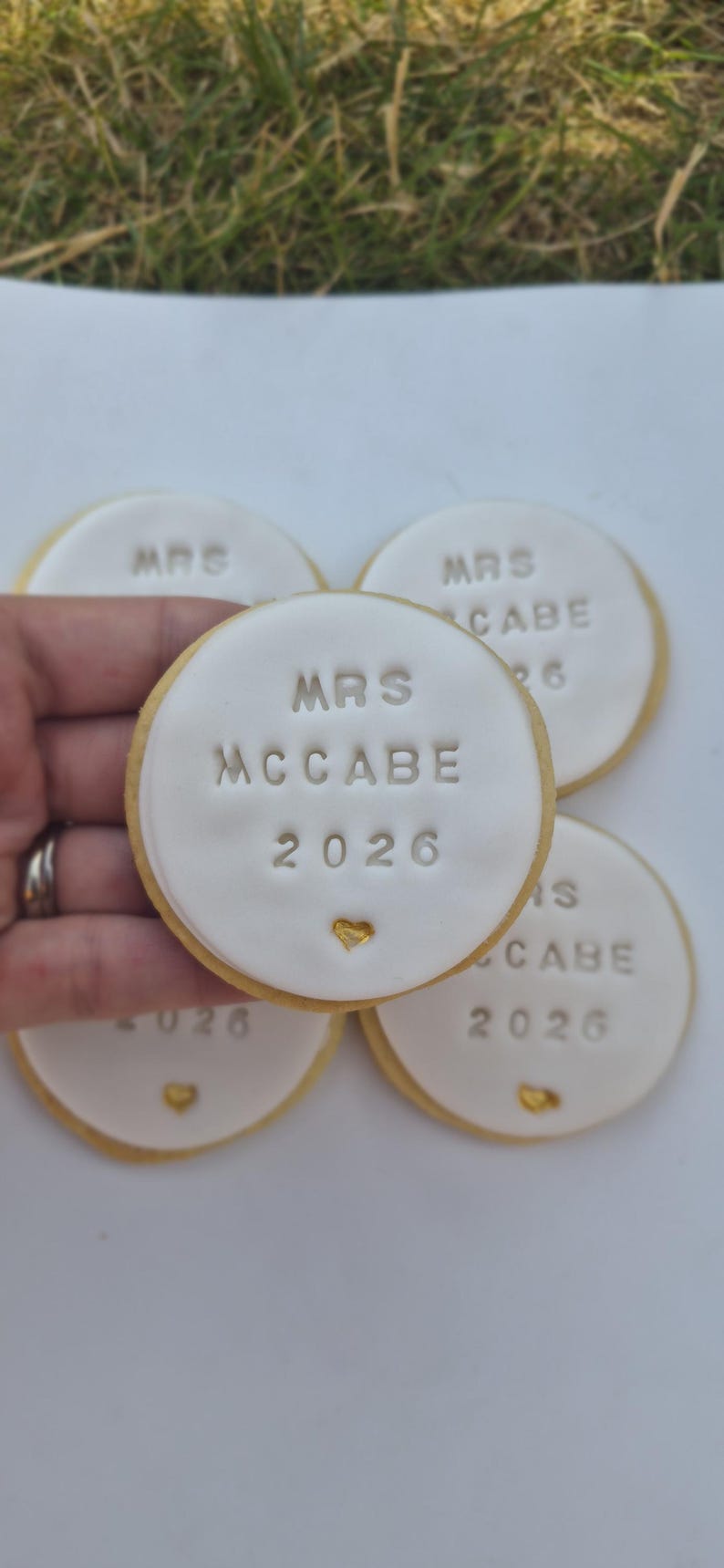 Puede incluir: Cuatro galletas de az&uacute;car redondas con glaseado blanco, cada una grabada con el texto MRS MCCABE 2026 y un peque&ntilde;o coraz&oacute;n dorado. Las galletas tienen un borde dorado claro y est&aacute;n dispuestas en grupo, con una sostenida en primer plano.
