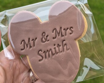 Op de afbeelding: Een koekje in de vorm van Mickey Mouse oren, bedekt met een bruine chocoladelaag. De woorden "Mr & Mrs Smith" zijn in de chocolade gestempeld. Het koekje zit in een transparante plastic verpakking.