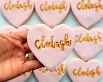 Op de afbeelding: Roze hartvormige koekjes met gouden letters die op elk koekje "Clodagh" zeggen.