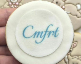 Op de afbeelding: Een rond wit koekje met de blauwe tekst Cmfrt in het midden gedrukt. Het koekje heeft een glad, plat oppervlak en is omringd door andere ronde koekjes versierd met reliëfafbeeldingen van een teddybeer, een konijn en een hert.