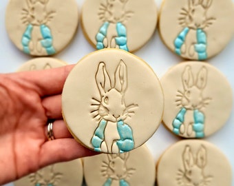 Op de afbeelding: Een set van negen gedecoreerde suikerkoekjes met een konijn dat een blauwe sjaal draagt. De koekjes zijn rond en hebben een lichtbruine kleur.