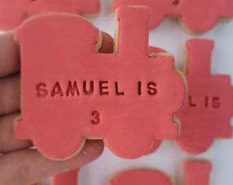 Op de afbeelding: Vijf rode, treinvormige suikerkoekjes versierd met witte glazuur en de tekst "Samuel is 3" op elk koekje.