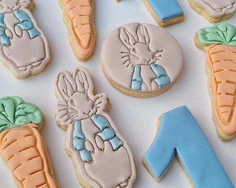 Op de afbeelding: Assortiment van versierde koekjes met Peter Konijn, wortels en het cijfer 1. De koekjes zijn in verschillende vormen en kleuren, waaronder roze, oranje, blauw en groen. De Peter Konijn koekjes hebben blauwe accenten.