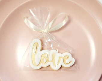 Op de afbeelding: Een witte koek met het woord "love" geschreven in gouden glazuur, verpakt in een doorzichtige plastic zak met een wit lint.