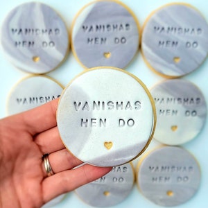 Puede incluir: Galletas redondas con un acabado de glaseado marmolado en gris y blanco. Cada galleta est&aacute; grabada con el texto VANISHAS HEN DO y presenta un peque&ntilde;o detalle de coraz&oacute;n dorado en la parte inferior. Tienen una superficie lisa y plana.