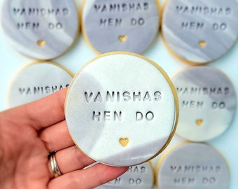 Op de afbeelding: Ronde koekjes met een gemarmerde grijs en witte glazuurlaag. Elk koekje is voorzien van de tekst VANISHAS HEN DO en heeft een klein gouden hartje aan de onderkant. De koekjes hebben een glad, plat oppervlak en een cirkelvorm.