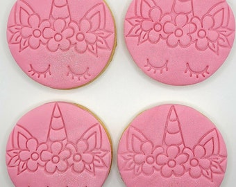 Op de afbeelding: Vier roze suikerkoekjes versierd met een eenhoornontwerp. Elk koekje heeft een eenhoornhoorn, gesloten ogen en een bloemenkrans.