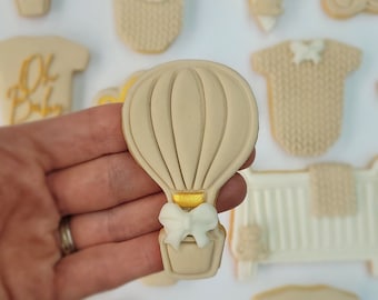 Op de afbeelding: Een set beige en witte gedecoreerde suikerkoekjes met verschillende babythema's, waaronder een luchtballon, een wieg, een rompertje en de woorden "Oh Baby" en "Baby".