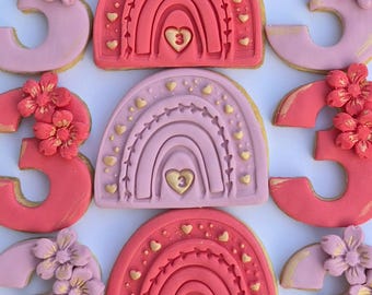 Op de afbeelding: Koekjes versierd met roze en rode glazuur. De koekjes hebben de vorm van regenbogen en het cijfer 3, met bloemen- en hartversieringen. De regenboogkoekjes hebben een hart met het cijfer 3 in het midden.