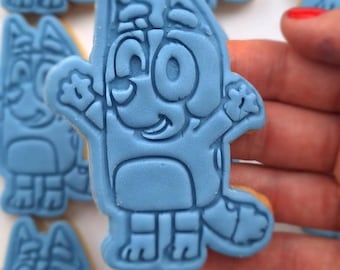 Op de afbeelding: Een lichtblauw koekje versierd met een verhoogd, reliëf ontwerp van een stripfiguurhond met grote oren, een brede glimlach en armen omhoog in een vrolijke houding, tegen een achtergrond van identieke blauwe koekjes.
