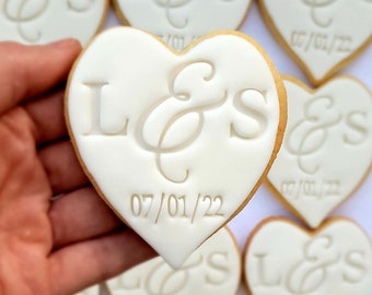Op de afbeelding: Een hartvormig koekje met glad wit glazuur, voorzien van de in reliëf aangebrachte letters L & S in het midden en de datum 07/01/22 eronder. Het koekje heeft een licht goudbruine rand en maakt deel uit van een groep identieke koekjes.