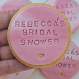 Puede incluir: Una galleta redonda cubierta con fondant rosa liso que presenta el texto en relieve REBECCA'S BRIDAL SHOWER y un peque&ntilde;o icono de coraz&oacute;n pintado en dorado en la parte inferior. La galleta tiene un borde dorado claro y textura marmolada.