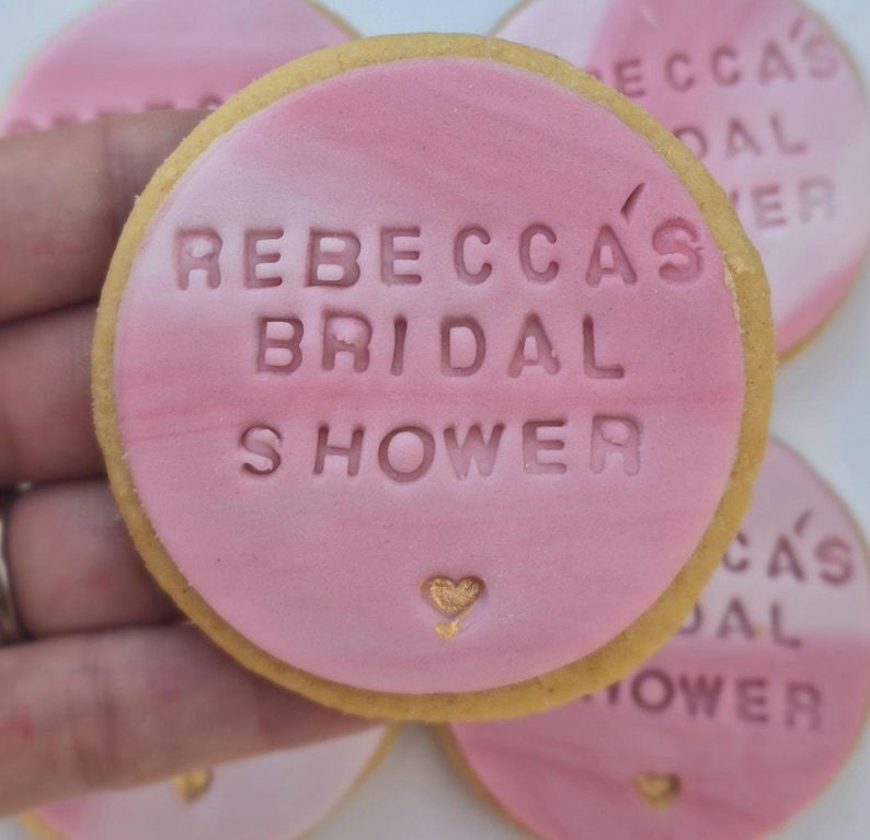 Puede incluir: Una galleta redonda cubierta con fondant rosa liso que presenta el texto en relieve REBECCA'S BRIDAL SHOWER y un peque&ntilde;o icono de coraz&oacute;n pintado en dorado en la parte inferior. La galleta tiene un borde dorado claro y textura marmolada.