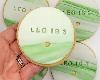 Op de afbeelding: Vier ronde suikerkoekjes versierd met groene en witte marmering. Elk koekje heeft een gouden hartje en de tekst "Leo is 3" in groene glazuur.