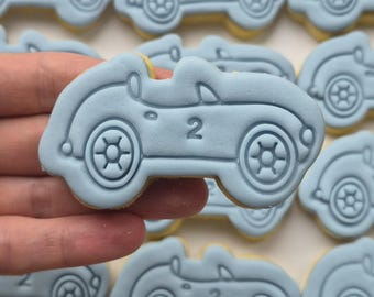 Op de afbeelding: Een verzameling lichtblauwe suikerkoekjes in de vorm van vintage cabriolets. Elk koekje heeft een reliëfontwerp van een auto met een duidelijk zichtbaar cijfer 2 op het zijpaneel, gedetailleerde wielen en een omlijning van de voorruit.