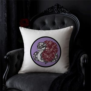 Może przedstawiać: Biała kwadratowa poduszka z kresk&oacute;wkowym wężem i kwiatowym wzorem. Wąż jest szary i biały, w czarnej obw&oacute;dce na fioletowym tle. Kwiaty są w głębokim bordowym kolorze. Poduszka leży na czarnym krześle.