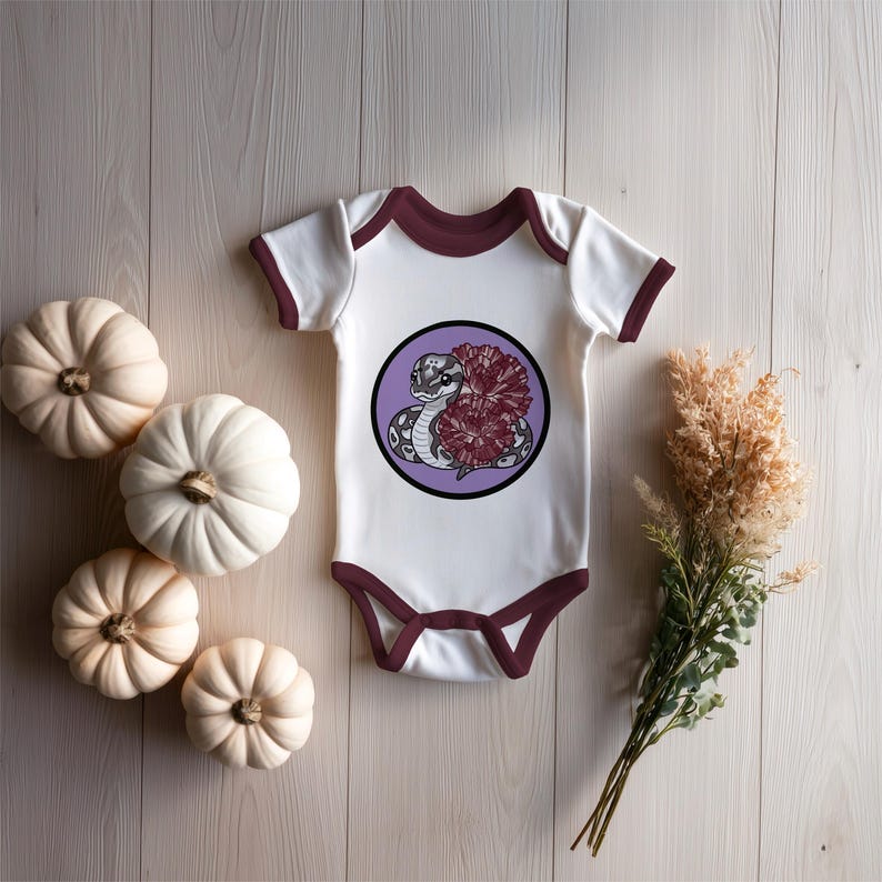 Może przedstawiać: Białe body dla dziecka z bordowymi wykończeniami, z okrągłą grafiką węża i kwiat&oacute;w na fioletowym tle. Body otoczone jest białymi dyniami i suszonymi kwiatami, tworząc jesienny motyw.