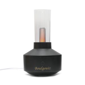 Puede incluir: Un difusor cilíndrico negro con una parte superior de cristal transparente. La base presenta el texto Boulgouiz en letras doradas sobre un botón de encendido. Dentro del cristal, un elemento de luz naranja central imita una llama. Un cable blanco.