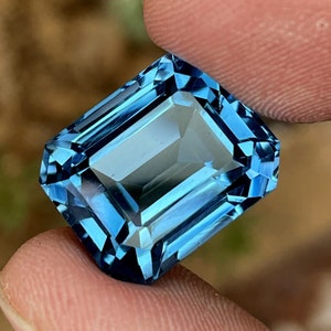 May include: A rectangular-cut gemstone in a deep blue shade. The gem is held between two fingers, highlighting its clarity and sparkle. The cut of the stone creates multiple facets, reflecting light. A beautiful piece of jewellery, perfect for a ring or pendant.