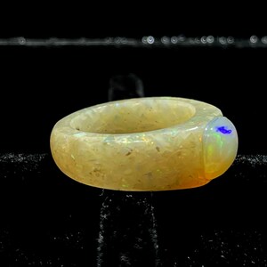 May include: A chunky, light-colored ring with a rounded shape. The ring has a speckled appearance with iridescent flecks. The gemstone on the ring's side has a blue and green color. Jewelry.