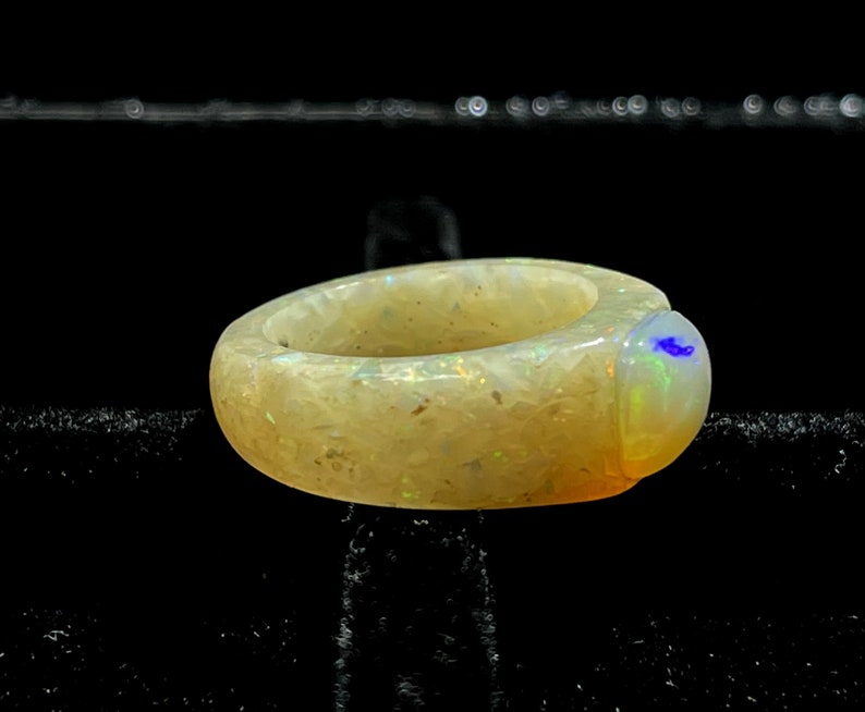 May include: A chunky, light-colored ring with a rounded shape. The ring has a speckled appearance with iridescent flecks. The gemstone on the ring's side has a blue and green color. Jewelry.