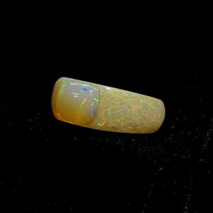 May include: A polished opal gemstone with a rectangular shape and rounded edges. The opal displays a creamy white base color with iridescent flashes of green, blue, and orange. The gemstone is set against a black background.