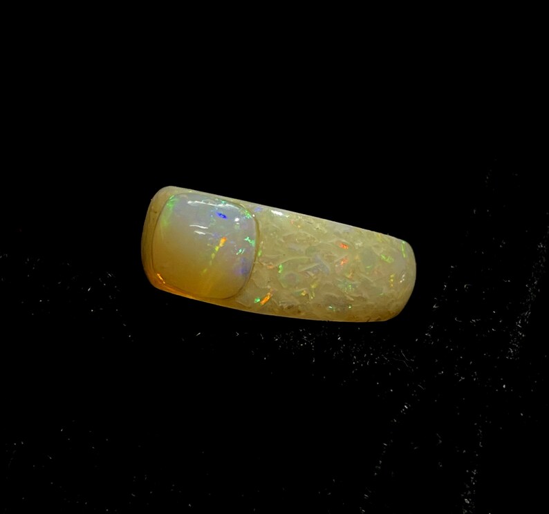 May include: A polished opal gemstone with a rectangular shape and rounded edges. The opal displays a creamy white base color with iridescent flashes of green, blue, and orange. The gemstone is set against a black background.