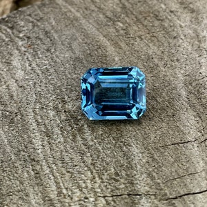 May include: A rectangular, faceted gemstone in a vibrant, light blue colour. The gem is set against a weathered, grey wooden surface, showcasing its clarity and brilliance. The cut of the stone is an emerald cut.