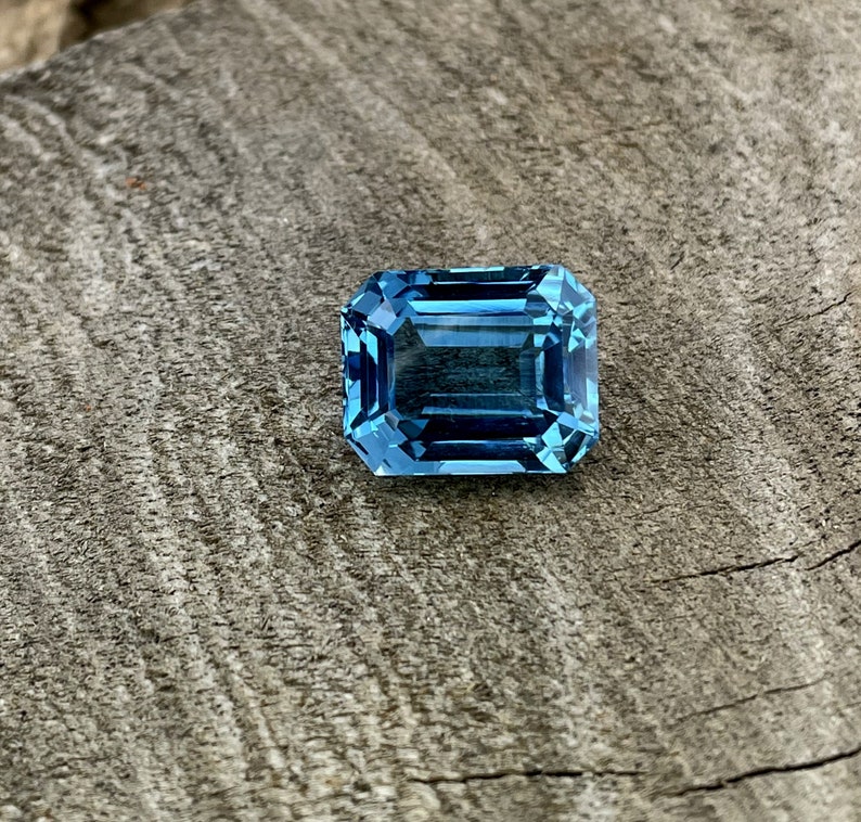 May include: A rectangular, faceted gemstone in a vibrant, light blue colour. The gem is set against a weathered, grey wooden surface, showcasing its clarity and brilliance. The cut of the stone is an emerald cut.