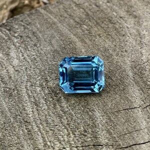 May include: A rectangular, light blue gemstone with an emerald cut, set against a weathered, grey wooden surface. The facets of the gem reflect light, creating a sparkling effect. The stone's colour is a vivid, clear blue, reminiscent of the sea.